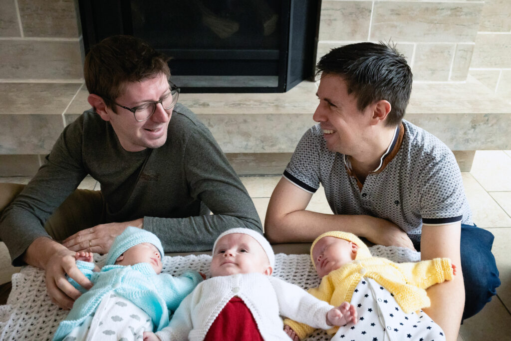 two dads looking at each other and smiling while holding their three babies